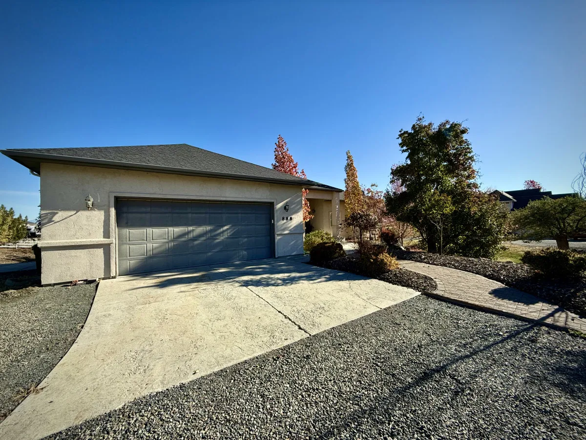 A beautifully landscaped exterior view with a garage door, a lush garden, and a stone pathway leading toward the home. The space exudes tranquility, with greenery framing the house and a sense of openness that hints at the surrounding scenery.