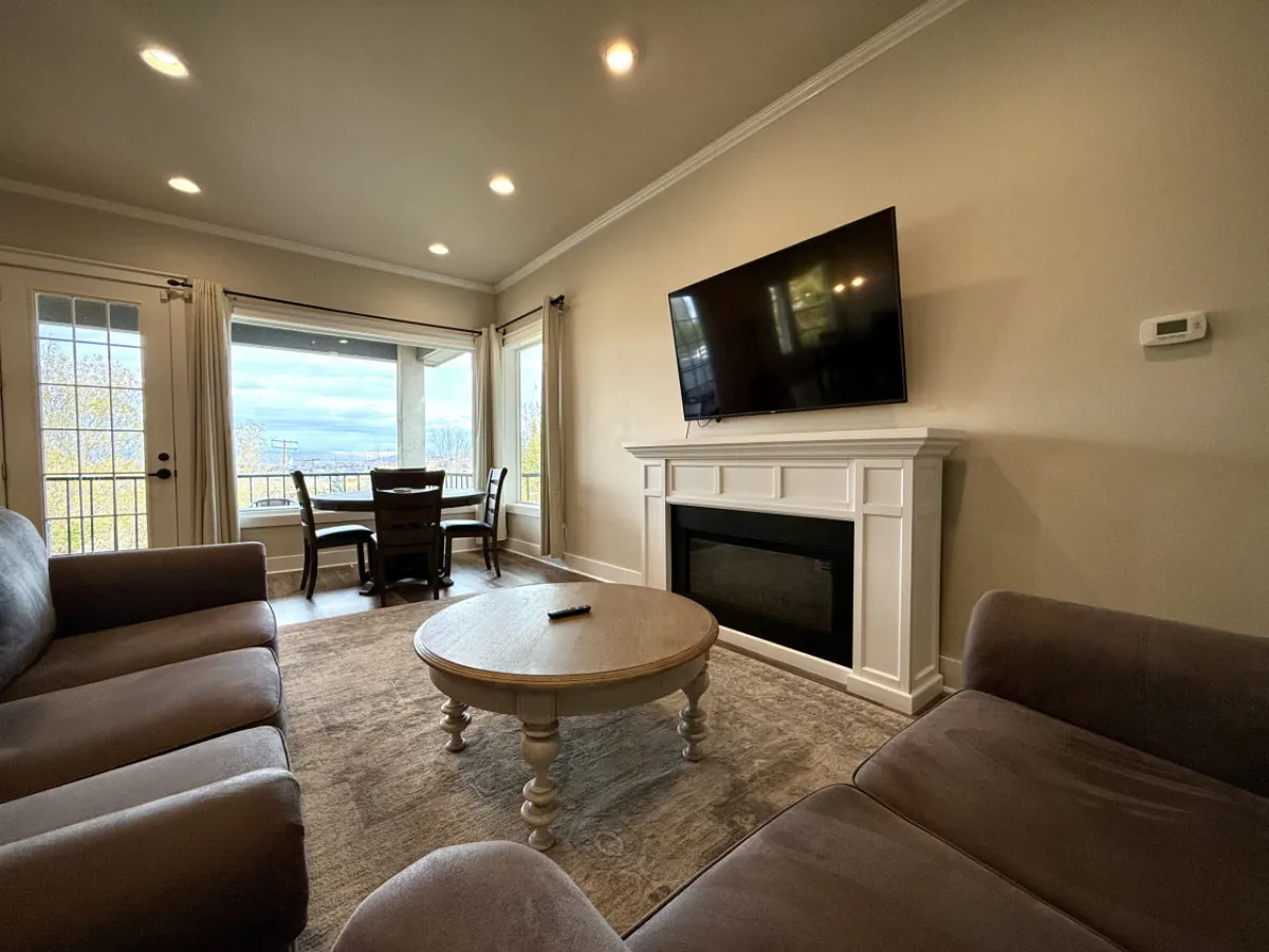 This image features an open-concept living and dining space, anchored by a plush sofa and coffee table facing a television and an inviting electric fireplace. The dining area with chairs is seamlessly visible, and ample natural light pours in through large windows, emphasizing the spacious and welcoming atmosphere.