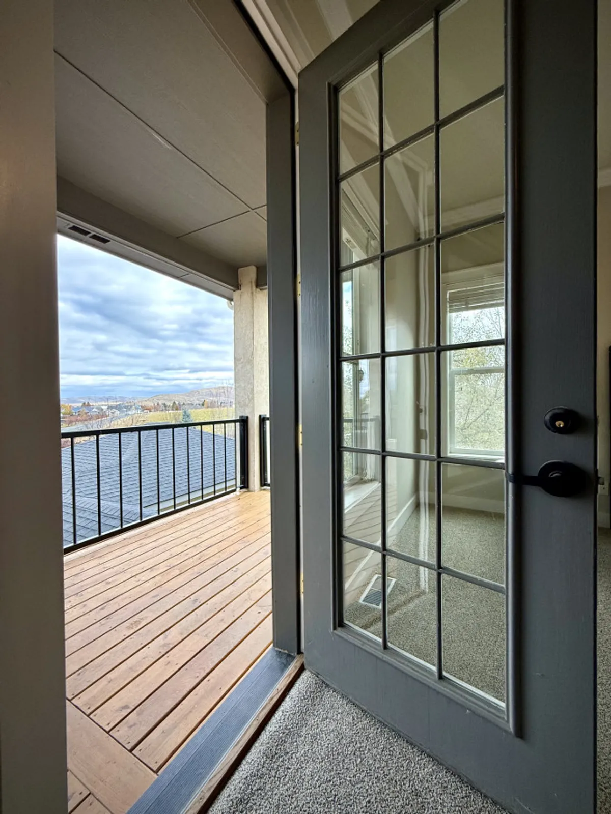 A view from inside looking out through double glass doors onto a wooden balcony overlooking a sweeping landscape. The inviting living area transitions seamlessly to the outdoors, with sunlight streaming in and the promise of sunsets visible from the deck.