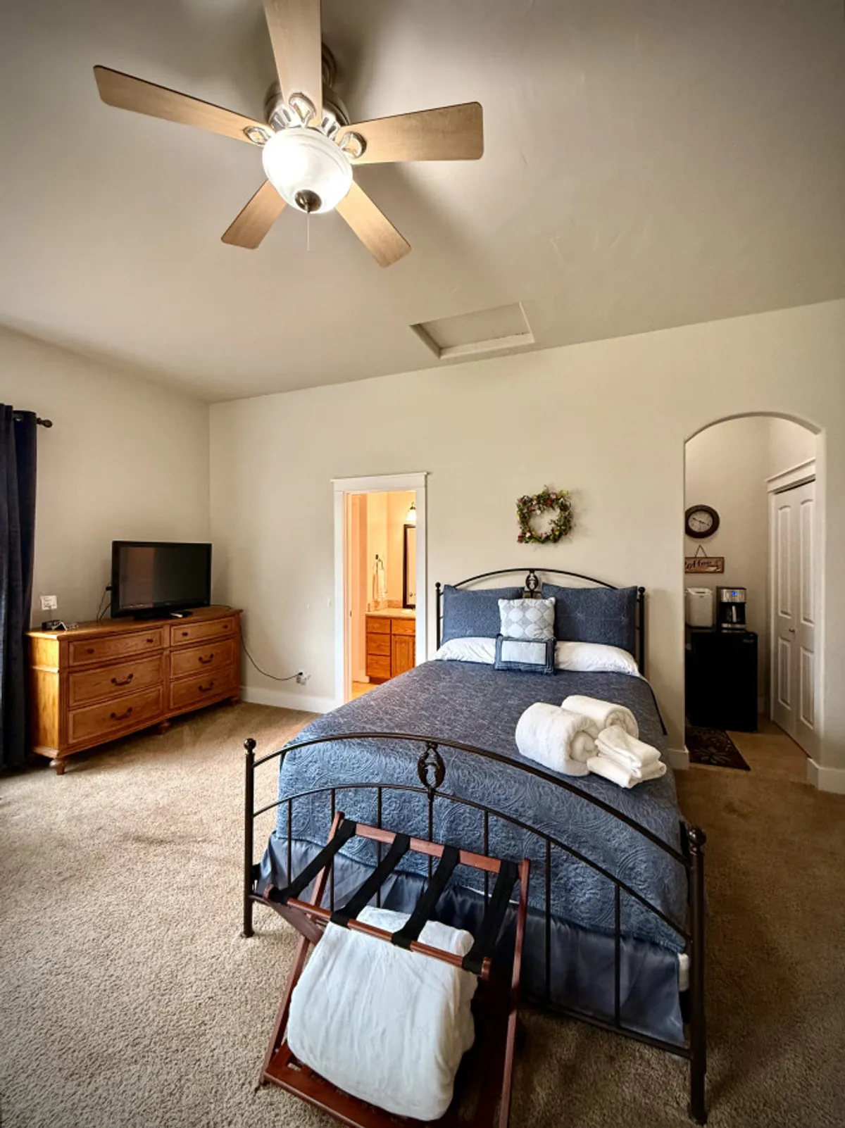 A serene bedroom with blue bedding, a wooden dresser, a television, and a decorative wreath on the wall. Sunlight streams through the window, illuminating the cozy space, ideal for rest and relaxation.