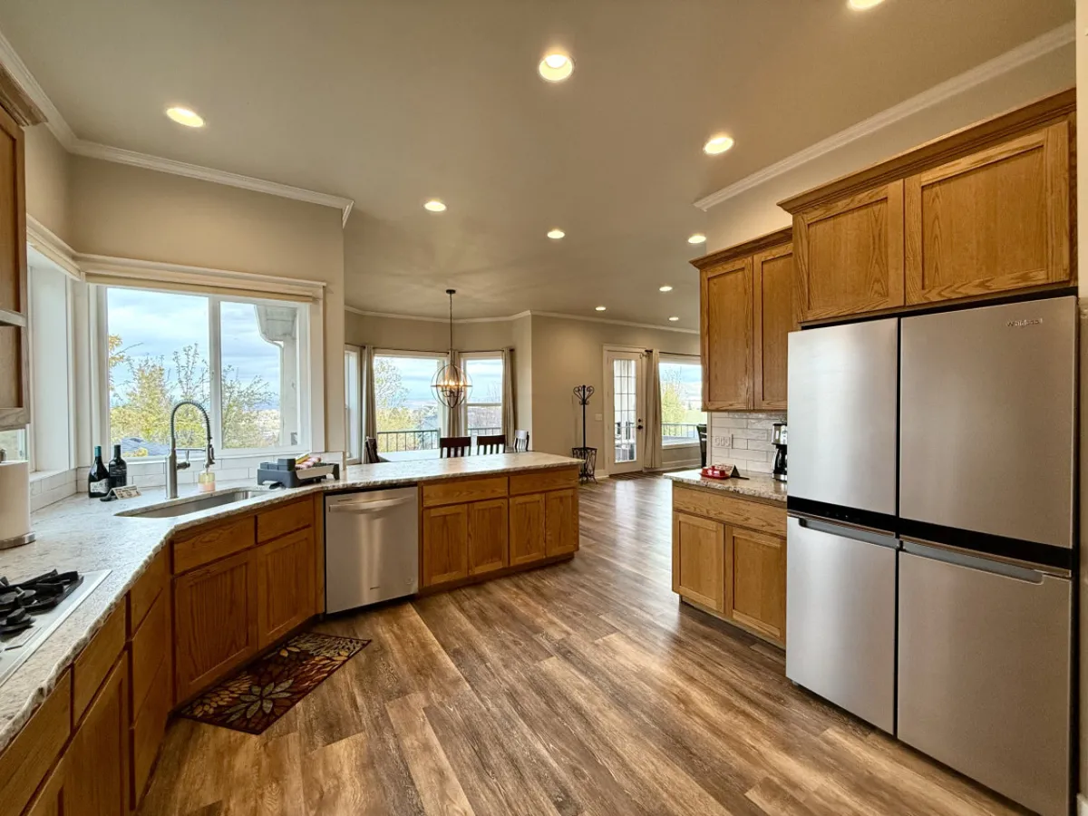 A spacious kitchen featuring a large central island, stainless steel appliances, and warm wooden cabinetry. The space is flooded with natural light from expansive windows, creating an inviting atmosphere ideal for both cooking and gathering. Bar seating lines the island, perfect for casual meals or lively tastings.